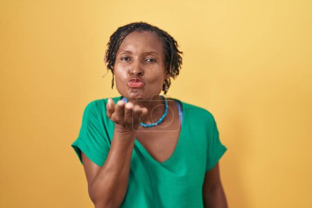 Téléchargez les photos : Femme africaine avec dreadlocks debout sur fond jaune regardant la caméra souffler un baiser avec la main sur l'air étant belle et sexy. expression d'amour. - en image libre de droit