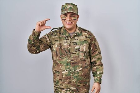 Foto de Joven hispano vistiendo uniforme camuflaje del ejército sonriente y confiado gesto con la mano haciendo letrero de pequeño tamaño con los dedos mirando y la cámara. concepto de medida. - Imagen libre de derechos