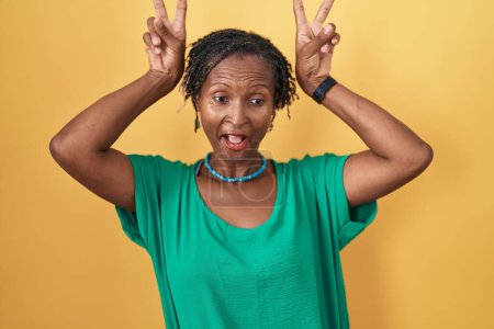 Foto de Mujer africana con rastas de pie sobre fondo amarillo posando divertido y loco con los dedos en la cabeza como orejas de conejo, sonriente alegre - Imagen libre de derechos