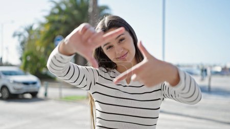 Foto de Joven hermosa mujer hispana sonriendo confiada haciendo gesto de marco con las manos en la calle - Imagen libre de derechos
