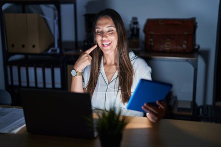 Téléchargez les photos : Jeune femme brune travaillant au bureau la nuit souriant joyeuse montrant et pointant du doigt les dents et la bouche. concept de santé dentaire. - en image libre de droit