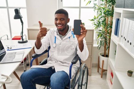 Téléchargez les photos : Afro-américain médecin homme assis sur fauteuil roulant tenant smartphone souriant heureux et positif, pouce levé faire excellent et signe d'approbation - en image libre de droit