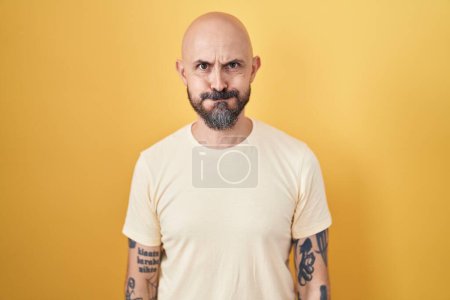 Téléchargez les photos : Homme hispanique avec des tatouages debout sur fond jaune joues gonflées avec drôle de visage. bouche gonflée d'air, expression folle. - en image libre de droit
