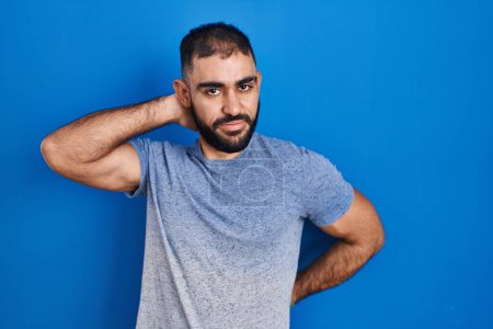 Téléchargez les photos : Moyen-Orient homme avec barbe debout sur fond bleu souffrant d'une blessure au cou, toucher le cou avec la main, douleur musculaire - en image libre de droit