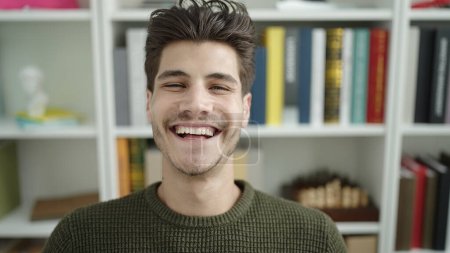 Téléchargez les photos : Jeune homme hispanique étudiant souriant confiant assis sur la table à l'université de la bibliothèque - en image libre de droit