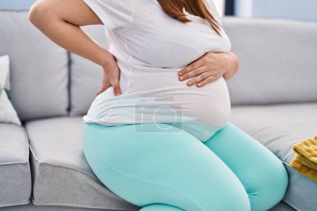Foto de Mujer embarazada joven que sufre de dolor de espalda sentado en el sofá en casa - Imagen libre de derechos
