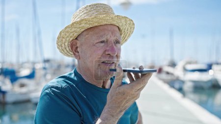 Foto de Senior hombre de pelo gris turista con sombrero de verano enviar mensaje de voz por teléfono inteligente en el puerto - Imagen libre de derechos