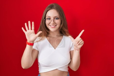 Téléchargez les photos : Jeune femme caucasienne debout sur fond rouge montrant et pointant vers le haut avec les doigts numéro sept tout en souriant confiant et heureux. - en image libre de droit