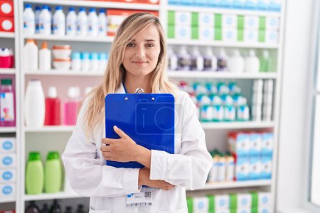 Téléchargez les photos : Jeune femme blonde pharmacien souriant confiant tenant presse-papiers à la pharmacie - en image libre de droit