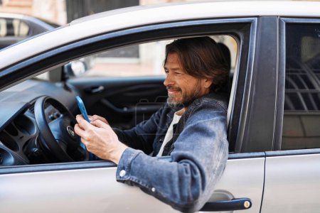 Téléchargez les photos : Homme d'âge moyen en utilisant smartphone assis sur la voiture à la rue - en image libre de droit