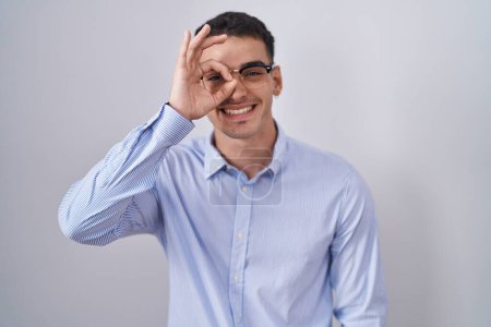 Foto de Hombre hispano guapo usando ropa de negocios y gafas haciendo buen gesto con la mano sonriente, los ojos mirando a través de los dedos con la cara feliz. - Imagen libre de derechos