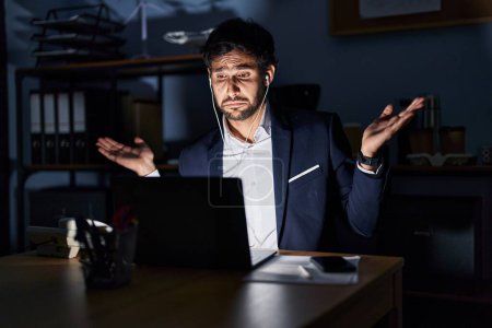 Photo for Handsome latin man working at the office at night clueless and confused expression with arms and hands raised. doubt concept. - Royalty Free Image