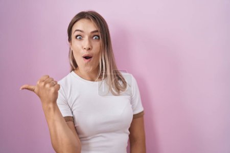 Photo for Blonde caucasian woman standing over pink background surprised pointing with hand finger to the side, open mouth amazed expression. - Royalty Free Image