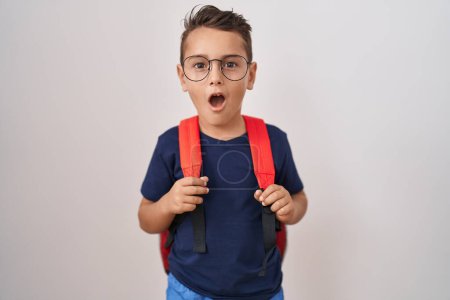 Foto de Pequeño niño hispano con gafas y mochila de estudiante asustado y sorprendido con la boca abierta para sorpresa, cara de incredulidad - Imagen libre de derechos