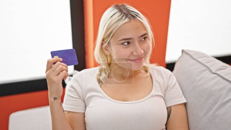 Téléchargez les photos : Jeune belle femme hispanique tenant la carte de crédit assis sur le canapé pensant à la maison - en image libre de droit