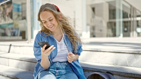 Téléchargez les photos : Jeune étudiante blonde utilisant un smartphone et un casque à l'université - en image libre de droit