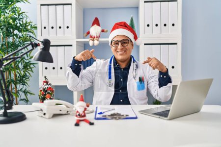 Foto de Joven médico hispano en la clínica en Navidad mirando confiado con sonrisa en la cara, señalándose con los dedos orgullosos y felices. - Imagen libre de derechos