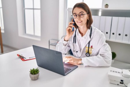 Photo for Young caucasian woman doctor using laptop talking on smartphone at clinic - Royalty Free Image