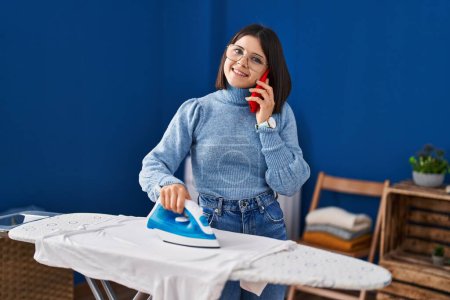 Photo for Young beautiful hispanic woman talking on smartphone ironing clothes at laundry room - Royalty Free Image