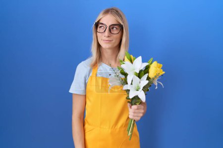 Téléchargez les photos : Jeune femme caucasienne portant tablier de fleuriste tenant des fleurs souriant regardant vers le côté et regardant loin en pensant. - en image libre de droit