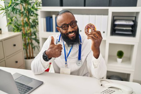 Foto de Hombre afroamericano trabajando en clínica dietista sosteniendo donut sonriendo feliz y positivo, pulgar hacia arriba haciendo excelente y signo de aprobación - Imagen libre de derechos