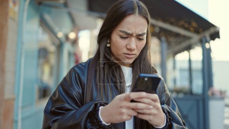 Téléchargez les photos : Jeune belle femme hispanique utilisant un smartphone avec une expression sérieuse sur la terrasse du café - en image libre de droit