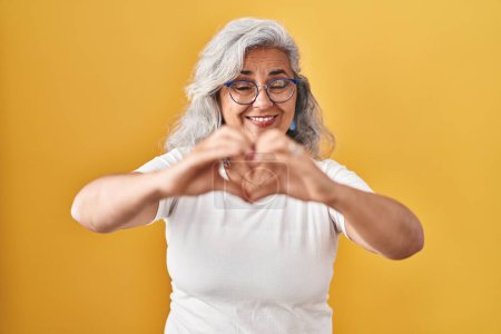 Photo for Middle age woman with grey hair standing over yellow background smiling in love doing heart symbol shape with hands. romantic concept. - Royalty Free Image