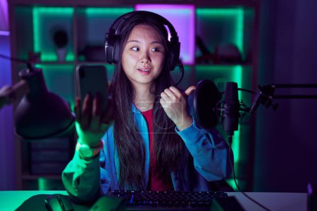Foto de Joven mujer asiática jugando videojuegos con smartphone sonriendo con cara feliz mirando y apuntando hacia un lado con el pulgar hacia arriba. - Imagen libre de derechos