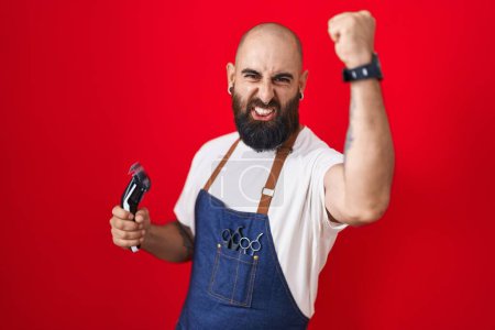 Photo for Young hispanic man with beard and tattoos wearing barber apron holding razor angry and mad raising fist frustrated and furious while shouting with anger. rage and aggressive concept. - Royalty Free Image