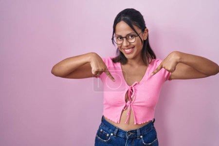 Photo for Hispanic young woman standing over pink background wearing glasses looking confident with smile on face, pointing oneself with fingers proud and happy. - Royalty Free Image