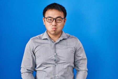 Téléchargez les photos : Jeune homme chinois debout sur fond bleu joues gonflées avec drôle de visage. bouche gonflée d'air, expression folle. - en image libre de droit