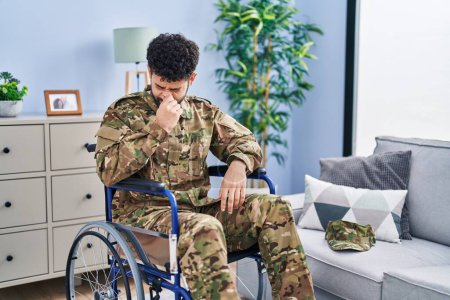 Photo for Arab man wearing camouflage army uniform sitting on wheelchair smelling something stinky and disgusting, intolerable smell, holding breath with fingers on nose. bad smell - Royalty Free Image