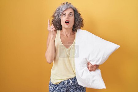 Téléchargez les photos : Femme d'âge moyen avec les cheveux gris portant un pyjama étreignant l'oreiller étonné et surpris regardant vers le haut et pointant avec les doigts et les bras levés. - en image libre de droit