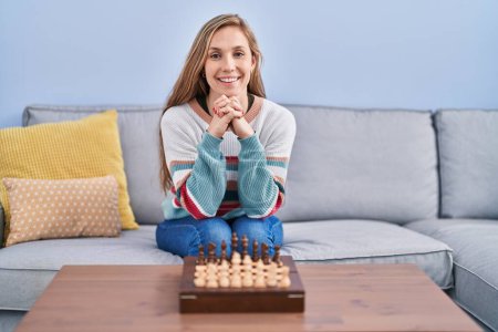 Téléchargez les photos : Jeune femme blonde jouant aux échecs assis sur le canapé regardant positif et heureux debout et souriant avec un sourire confiant montrant les dents - en image libre de droit