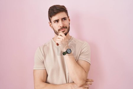Foto de Hombre hispano con barba de pie sobre fondo rosa pensando preocupado por una pregunta, preocupado y nervioso con la mano en la barbilla - Imagen libre de derechos