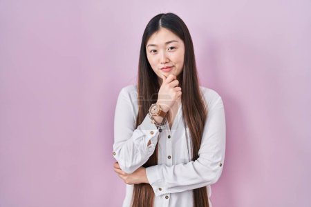Foto de Mujer joven china de pie sobre fondo rosa mirando con confianza a la cámara sonriendo con los brazos cruzados y la mano levantada en la barbilla. pensar positivamente. - Imagen libre de derechos