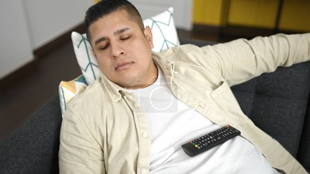 Téléchargez les photos : Jeune homme hispanique tenant télécommande tv couché sur le canapé dormant à la maison - en image libre de droit