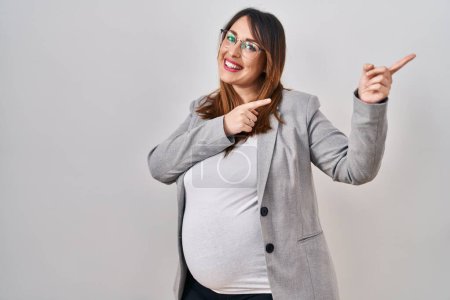 Foto de Embarazada mujer de negocios de pie sobre fondo blanco sonriendo y mirando a la cámara apuntando con dos manos y dedos a un lado. - Imagen libre de derechos