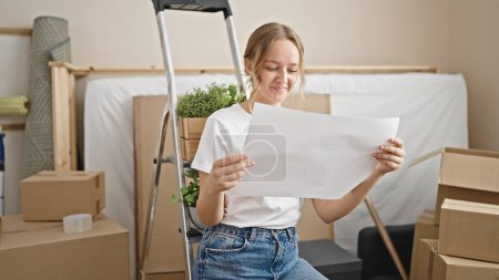 Photo for Young blonde woman sitting on ladder reading house project at new home - Royalty Free Image