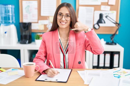 Foto de Young hispanic woman working at the office wearing glasses smiling doing phone gesture with hand and fingers like talking on the telephone. communicating concepts. - Imagen libre de derechos