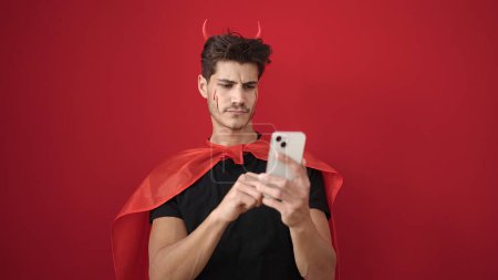 Foto de Joven hombre hispano usando disfraz de diablo usando teléfono inteligente sobre fondo rojo aislado - Imagen libre de derechos