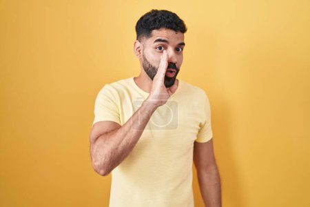 Téléchargez les photos : Homme hispanique avec barbe debout sur fond jaune main sur bouche racontant rumeur secrète, chuchotant conversation malicieuse - en image libre de droit