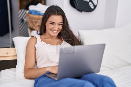 Téléchargez les photos : Jeune femme hispanique utilisant un ordinateur portable couché sur le lit dans la chambre - en image libre de droit
