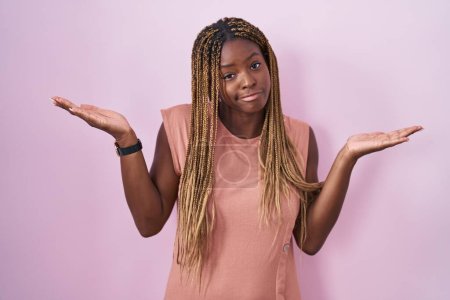 Foto de Mujer afroamericana con el pelo trenzado de pie sobre fondo rosa expresión despistada y confusa con los brazos y las manos levantadas. concepto de duda. - Imagen libre de derechos