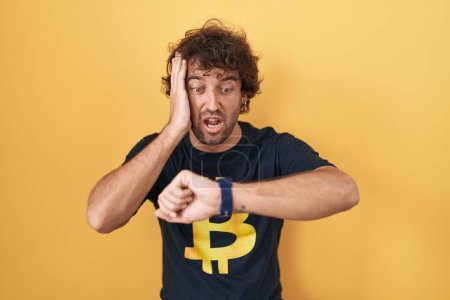 Téléchargez les photos : Jeune homme hispanique portant Bitcoin t-shirt regardant l'heure de la montre inquiet, peur de se faire tard - en image libre de droit