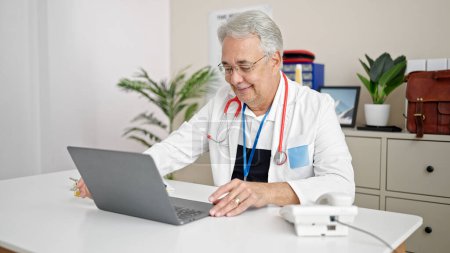 Téléchargez les photos : Homme d'âge moyen avec médecin des cheveux gris utilisant un ordinateur portable travaillant à la clinique - en image libre de droit