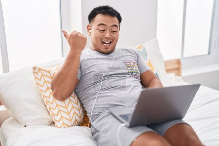 Téléchargez les photos : Chinois jeune homme à l'aide d'un ordinateur portable assis sur le lit pointant pouce vers le côté souriant heureux avec la bouche ouverte - en image libre de droit