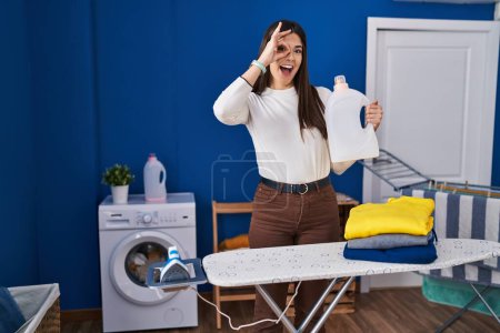 Photo for Young brunette woman with folded laundry after ironing smiling happy doing ok sign with hand on eye looking through fingers - Royalty Free Image