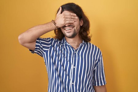 Téléchargez les photos : Homme hispanique avec les cheveux longs debout sur fond jaune souriant et riant avec la main sur le visage couvrant les yeux pour la surprise. concept aveugle. - en image libre de droit