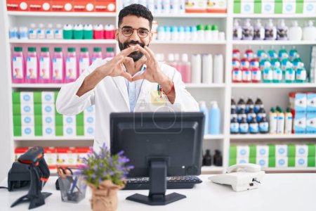 Téléchargez les photos : Homme hispanique avec barbe travaillant à la pharmacie pharmacie souriant dans l'amour faisant forme de symbole de coeur avec les mains. concept romantique. - en image libre de droit
